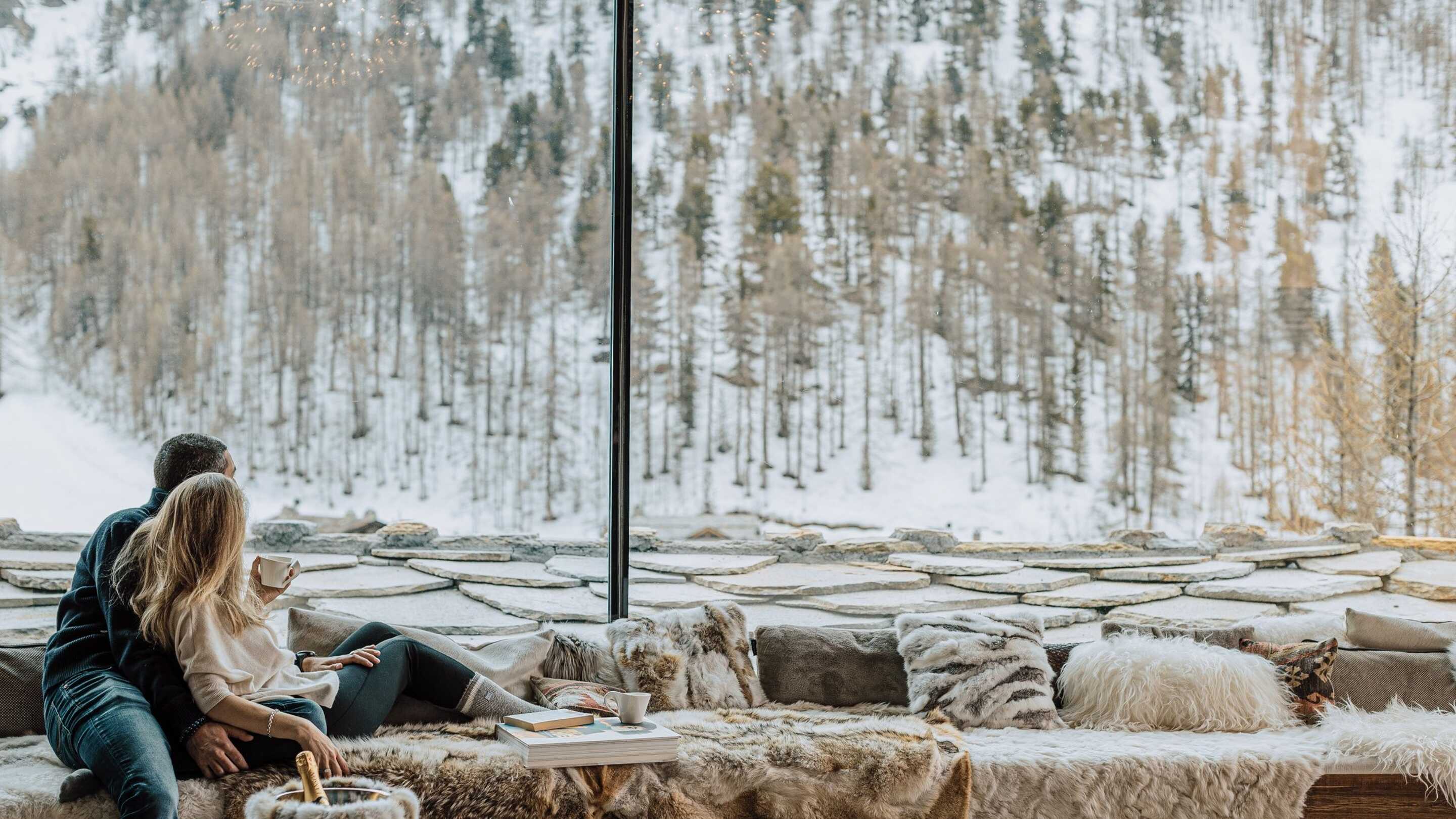 jeune-couple-regardant-une-vue-panoramique-sur-la-montagne-enneigee-depuis-la-baie-vitree-chalet-de-luxe-val-d-isere