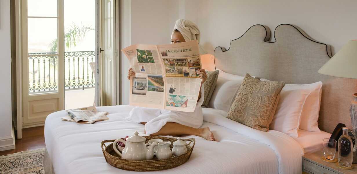 Woman-in-a-dressing-gown-reading-a-newspaper-on-a-bed-with-a-tray-of-tea